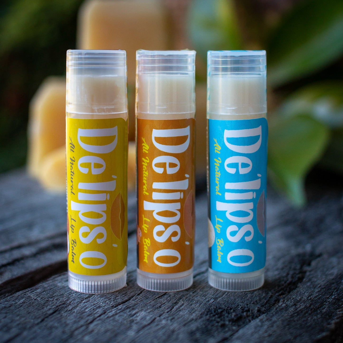 Three Delipso lip balms on a wooden surface with a blurred natural background.