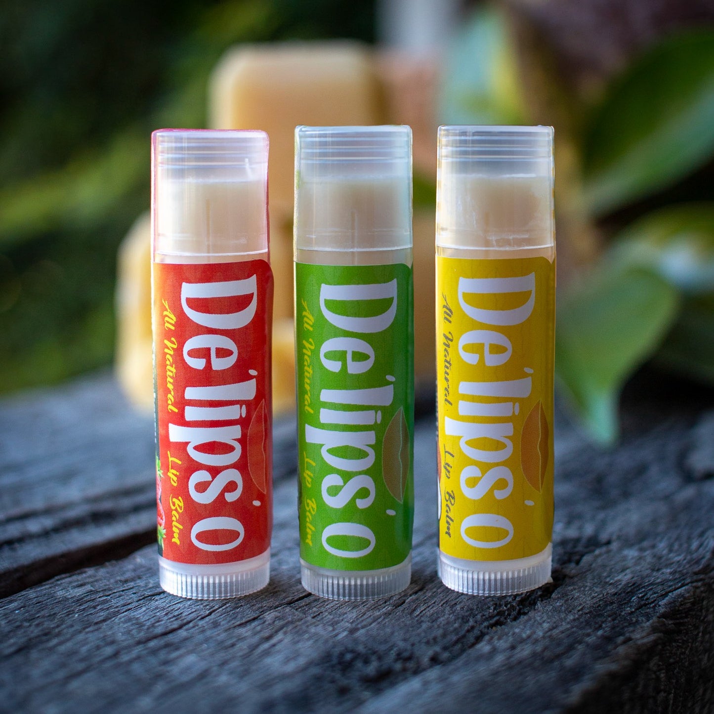 Three Delipso lip balms on a wooden surface with a blurred natural background.